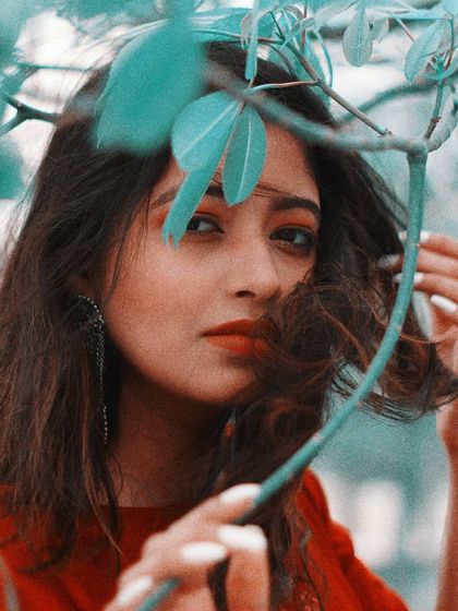 A close-up portrait with a creative twist, using leaves to frame the model's face. The teal and red color grading gives the image a unique, artistic feel while drawing attention to her direct gaze and the simple beauty of the natural elements.