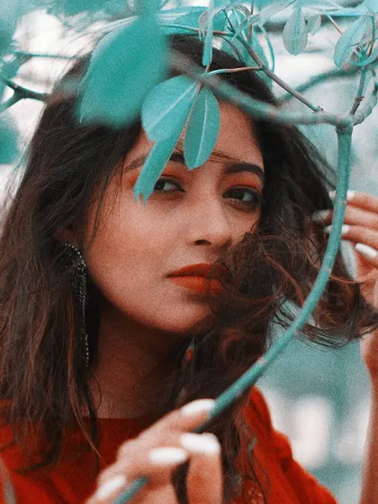 A close-up portrait with a creative twist, using leaves to frame the model's face. The teal and red color grading gives the image a unique, artistic feel while drawing attention to her direct gaze and the simple beauty of the natural elements.