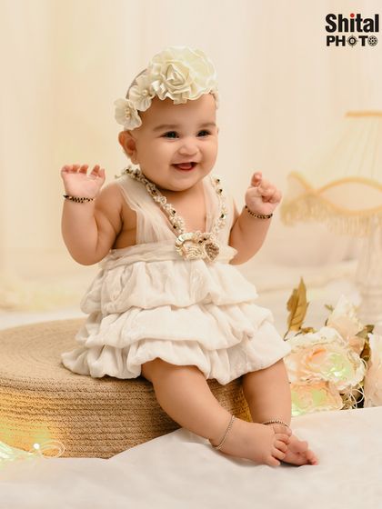 A full-length portrait of a baby girl in a tiered ruffle dress and floral headband, sitting on a rustic jute stool.
