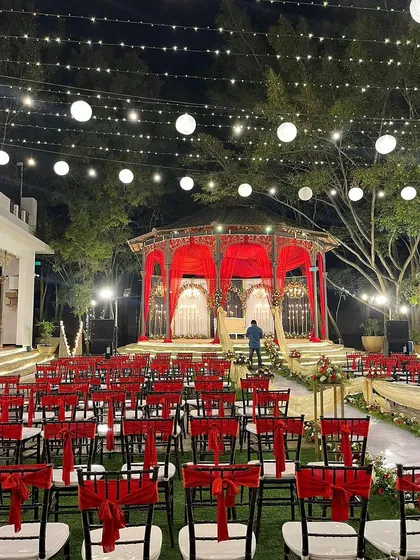 A beautiful outdoor reception setup with a red-draped gazebo stage and an aisle lined with flowers, all under a canopy of glowing lanterns.
