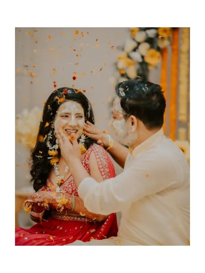 A sweet moment where the groom gently applies haldi to the bride's face, with flower petals falling around them.