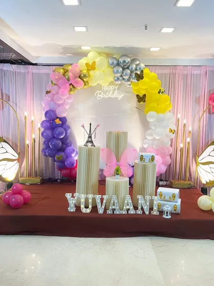 A beautiful butterfly themed birthday stage with a ring backdrop. The setup includes a multi-colored balloon garland, large illuminated butterfly wings, and a personalized name sign in light-up letters.