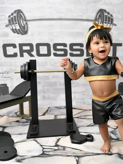 A triumphant smile from the strongest one-year-old in the studio. Holding her dumbbell high, she's clearly having a blast in her gym-themed session.