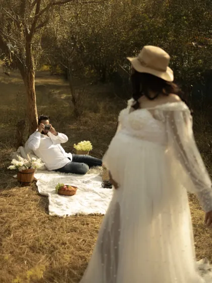 A creative, behind-the-scenes style shot. The mother-to-be is in the blurred foreground, while the focus is on her partner in the background, taking a photo of her, telling a story of mutual love and admiration.