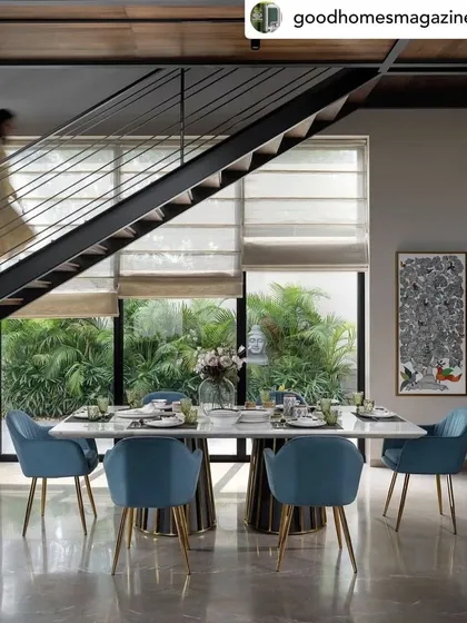 The dining area in the Dua Residence, set against a backdrop of lush greenery visible through floor-to-ceiling windows. The vibrant blue chairs add a beautiful pop of color to the otherwise neutral and serene space.