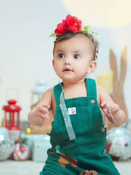 A baby girl in a green corduroy jumper enjoys her Christmas mini session, surrounded by festive lights and decorations.