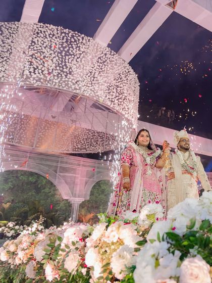 The couple stands proudly on stage, framed by an explosion of white flowers and celebratory cold pyrotechnics. This image captures the triumphant and joyous spirit of the moment.