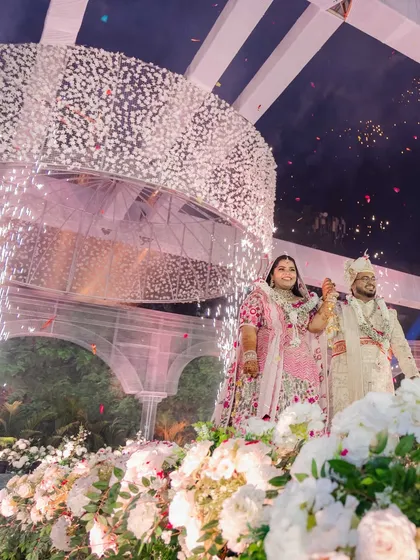 The couple's grand entrance, surrounded by sparklers and standing amidst a field of white flowers. The large floral chandelier above them adds to the drama and beauty of the moment.