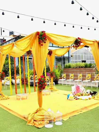 The central mandap for a Haldi ceremony, draped in bright yellow fabric and decorated with red roses and marigold strings.