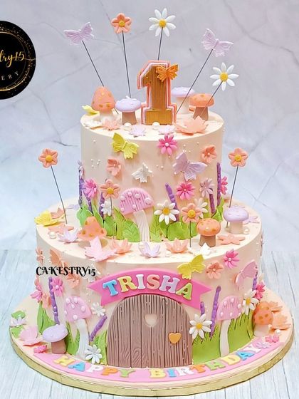 An enchanting two-tier chocolate cake with a fairy garden theme. The cake is covered in fondant flowers, mushrooms, and butterflies, with a whimsical door at the base.