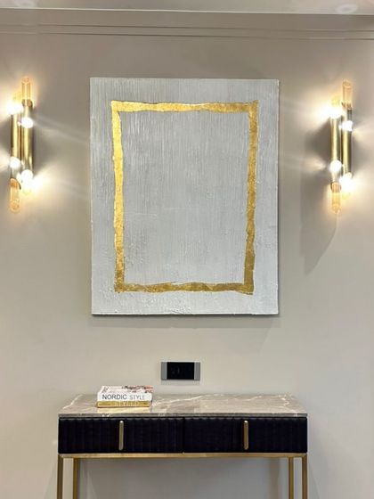 A beautiful art installation in a Hyderabad home. This piece features a simple, elegant design with a textured white background and a border of 24ct gold leaf, perfectly framed by the wall sconces.