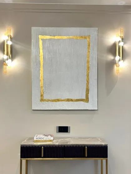 A beautiful art installation in a Hyderabad home. This piece features a simple, elegant design with a textured white background and a border of 24ct gold leaf, perfectly framed by the wall sconces.