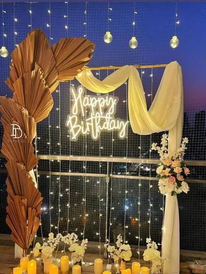 An elegant balcony decoration with a 'Happy Birthday' neon sign as the centerpiece. The setup is framed by dried palm leaves, draped fabric, fairy lights, and flameless candles, creating a magical evening ambiance.