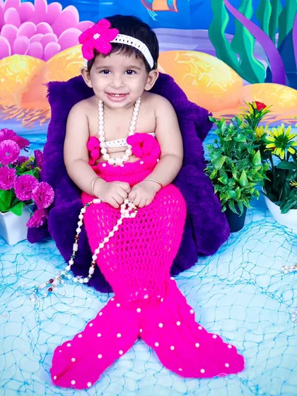A smiling baby girl in a pink mermaid costume, holding a string of pearls.