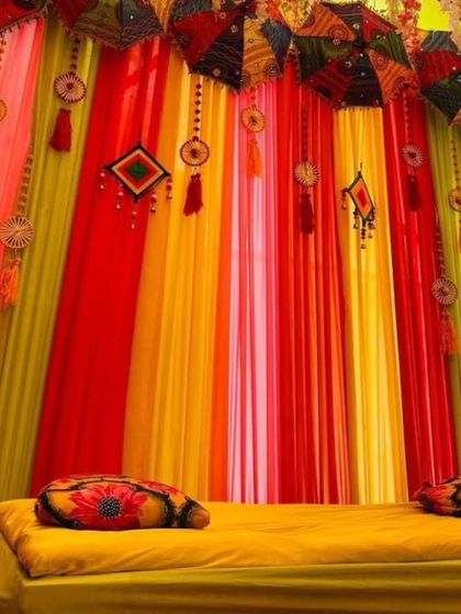 A close-up of a colorful Mehandi seating area. The decor features a low gadda with printed cushions and a backdrop of bright drapes with hanging Rajasthani umbrellas.