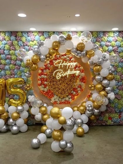 A luxurious 75th birthday party with a full floral wall. The setup is enhanced with a gold and silver balloon arch, a neon sign, and large '75' foil balloons.