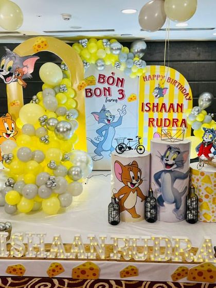 A playful Tom and Jerry theme party, with multiple character cutouts, cheese-themed details, and a yellow and silver balloon arrangement.