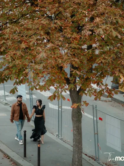 An observational shot of a couple walking down a Parisian sidewalk, framed by an autumn tree. This candid photo captures a simple, everyday moment made romantic by the city setting.