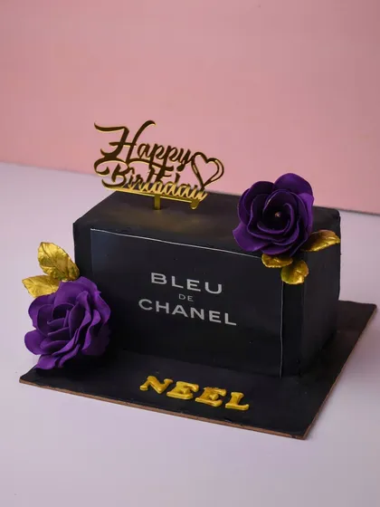 A chic cake designed to look like a Bleu de Chanel perfume bottle. This rectangular cake is covered in black fondant with silver lettering and adorned with deep purple sugar flowers and gold leaves for a touch of luxury.