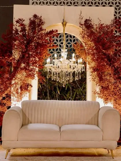 The reception stage, where a simple white sofa is set against a backdrop of arched panels, crystal chandeliers, and dramatic bursts of red foliage, creating a look of modern romance.