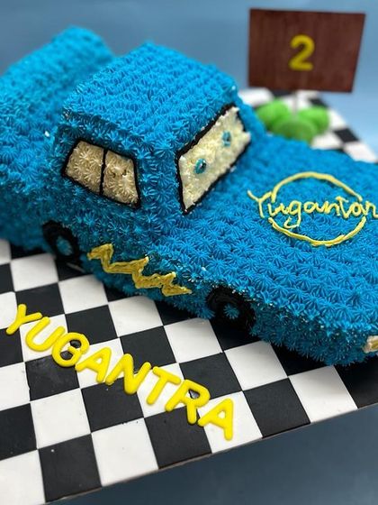 A custom-sculpted car cake for a second birthday. This cake is shaped like a blue race car and decorated with intricate buttercream piping to create a unique texture, complete with a checkered flag base.