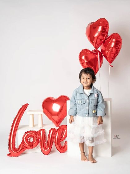A toddler photoshoot with a 'Love' theme, perfect for Valentine's Day or any day. She poses with red heart-shaped balloons and a denim jacket, creating a sweet and modern portrait.