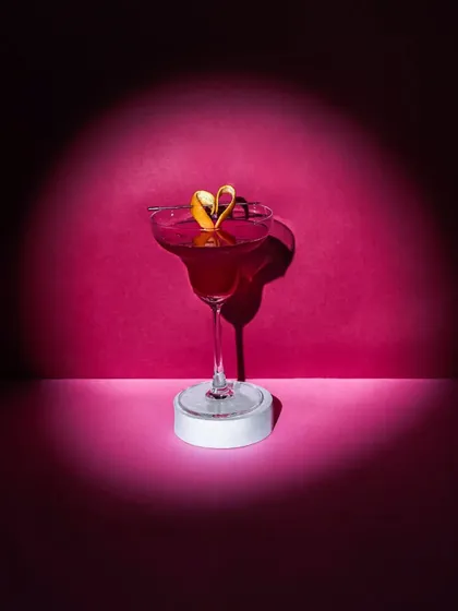 A single, elegant red cocktail in a margarita glass, highlighted by a dramatic spotlight against a dark pink background. The heart-shaped garnish makes it perfect for a Valentine's Day promotion.