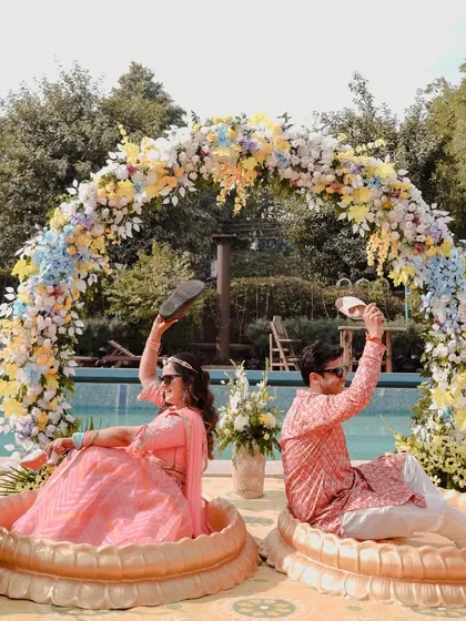 A playful Haldi portrait of the couple sitting in large decorative urns by the pool, holding up slippers in a fun, quirky pose that shows off their personalities.