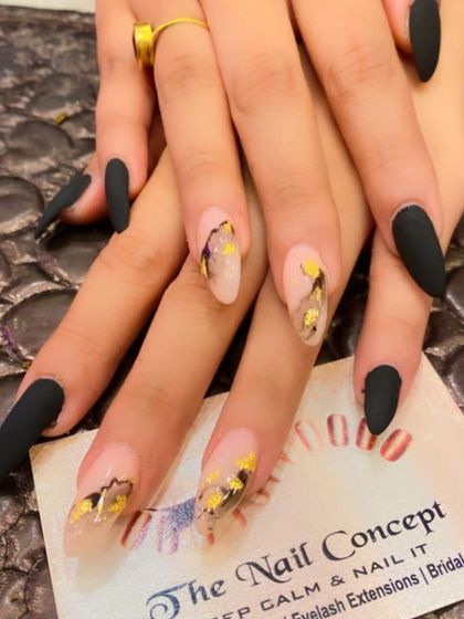 A dramatic and artistic set of long, matte black stiletto nails. The accent nails have a sheer base with an abstract design using black lines and gold foil flakes.