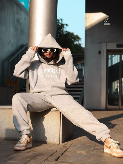 An urban streetwear shoot featuring a model in a grey tracksuit. The low angle and confident pose create a cool, edgy vibe against the concrete and metal background.