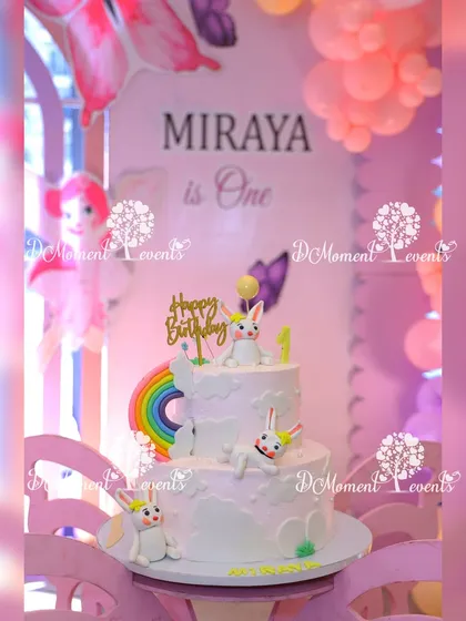 A close-up of a bunny-themed birthday cake. The two-tiered white cake is adorned with cute bunny figures and a small rainbow, perfectly matching the party's theme.