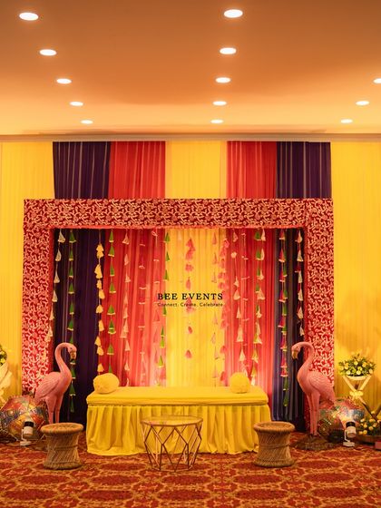 A colorful and quirky Mehendi stage. The backdrop features vibrant drapes in yellow, pink, and purple, with fun elements like flamingo props and tassel hangings, creating a lively and celebratory mood.
