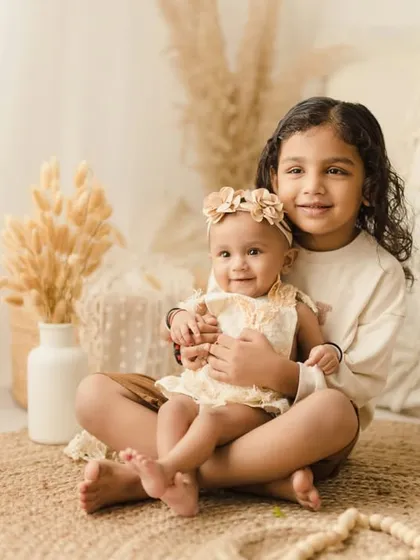 A sweet sibling portrait where the older sister holds her baby sister in her lap. The photo is set in our cozy boho studio with natural textures and tones.
