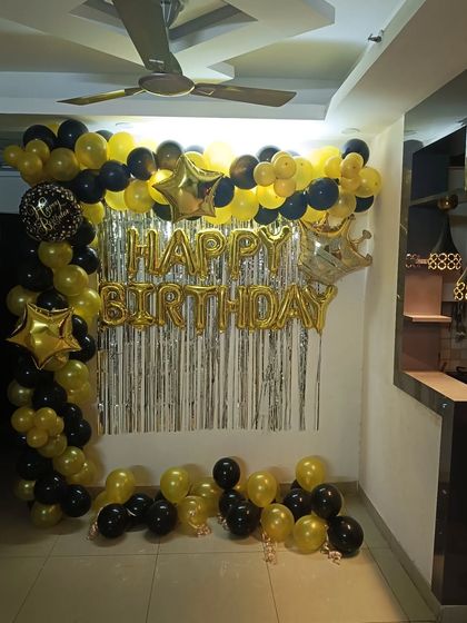 A black and gold birthday decoration for a home party. This setup includes a balloon arch, 'Happy Birthday' foil letters, and star-shaped foil balloons.