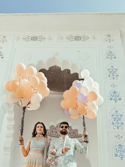 A creative low-angle shot of the couple holding balloons, framed by a beautiful white archway, capturing the fun carnival theme of their pre-wedding event.