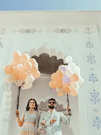 A creative low-angle shot of the couple holding balloons, framed by a beautiful white archway, capturing the fun carnival theme of their pre-wedding event.