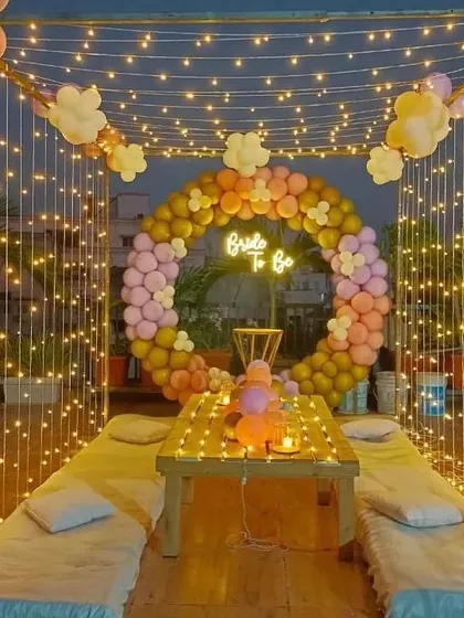 I created this magical bride-to-be party on a terrace, perfect for an evening celebration. The setup includes a canopy of fairy lights, low seating with cushions, and a circular balloon arch in pastel shades. A 'Bride to Be' neon sign serves as the centerpiece for photos.