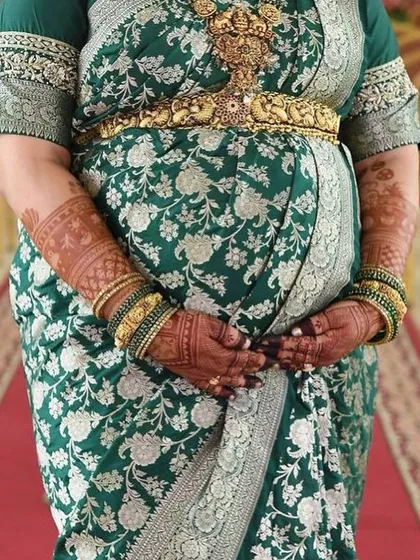 A close-up shot focusing on the baby bump and the beautifully stained mehendi. The deep reddish-brown color of the henna looks stunning against her traditional green and silver saree.