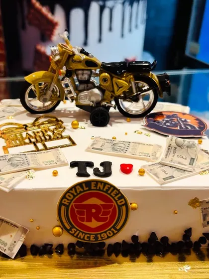 A Royal Enfield-themed cake featuring a miniature Bullet motorcycle, the brand's logo, and edible money notes.