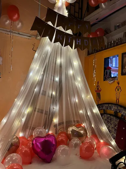 A cozy and romantic birthday surprise in a bedroom. The setup includes a canopy tent with fairy lights over the bed, surrounded by red and pink balloons.