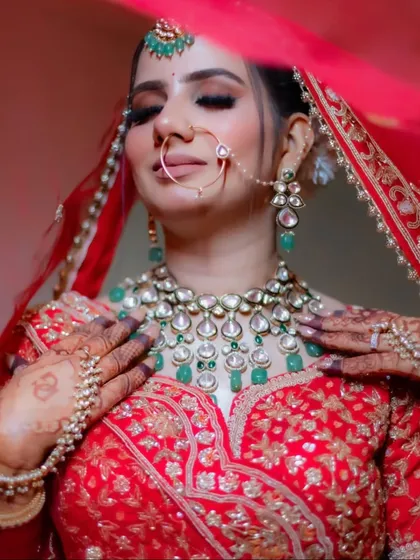 A close-up of the bride adjusting her stunning emerald and polki necklace, with the veil softly framing her face. This shot focuses on the luxurious bridal jewelry.
