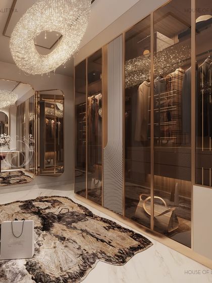 A view of a luxurious walk-in closet, where mirrored finishes, soft lighting, and curated detail are at every glance. The space includes glass-fronted wardrobes, a central island with a tufted ottoman, and a statement chandelier, celebrating the private joy of dressing up.
