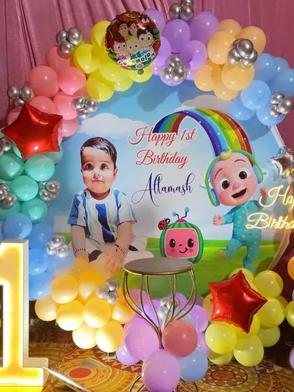 A colorful and fun Cocomelon theme decoration for a first birthday party. The setup includes a custom printed backdrop with the birthday boy's photo, a pastel balloon arch, a light-up marquee number '1', and Cocomelon character cutouts.