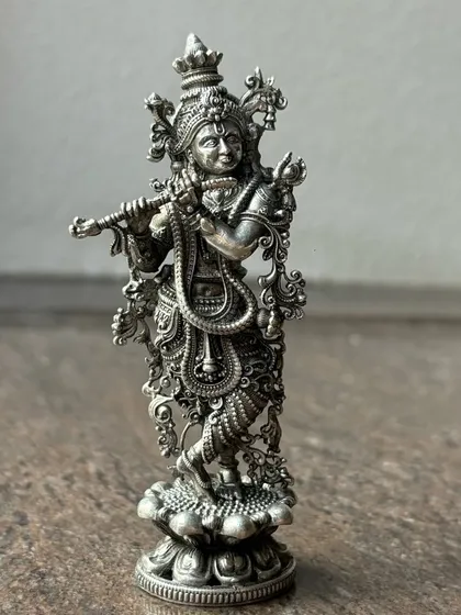 An intricately designed silver murti of Lord Krishna playing the flute. The oxidized finish enhances the detailed carvings on his attire and ornaments, giving it a timeless, antique look.