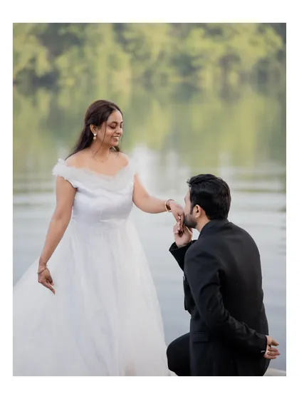 The groom-to-be kisses his fiancée's hand during their pre-wedding shoot by the water, a classic romantic gesture captured in beautiful natural light.