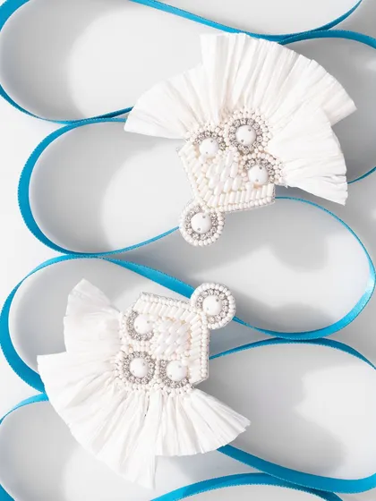 A clean and bright shot of white, fan-shaped earrings against a backdrop of curving blue ribbon. The composition is elegant and graphic, highlighting the intricate details of the jewelry.