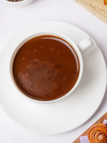 A close-up, top-down view of a cup of rich, dark hot chocolate. The minimalist styling on a white saucer makes the beverage itself the star of the shot.