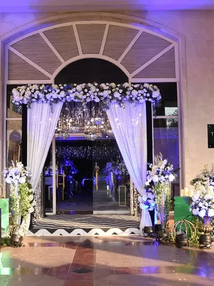 A chic and glamorous entrance for a Sangeet night, with white drapes, a canopy of flowers, and cool blue lighting setting a sophisticated party mood.