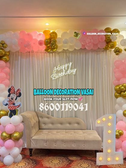 A classic Minnie Mouse theme for a first birthday party in a hall. The setup includes a pink, white, and gold balloon arch, a comfortable divan for photos, and a light-up number one.
