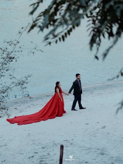 A wide shot of a couple walking hand-in-hand on a white sandy beach, with her red gown trailing behind. The framing through the trees adds depth.